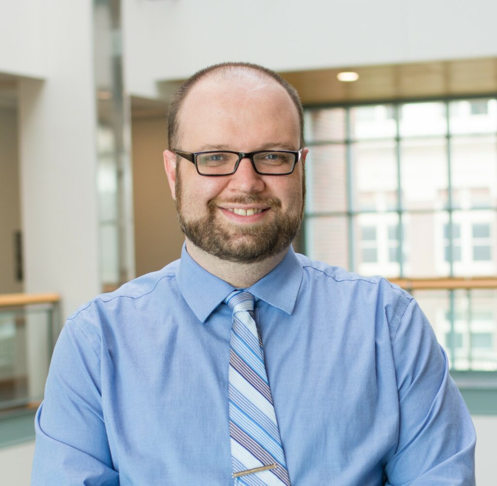 Head shot of Ian MacFarlane, a cis-man wearing a blue button up shirt with a blue tie. He has a short beard, black glasses, and short hair with a rapidly retreating hairline.